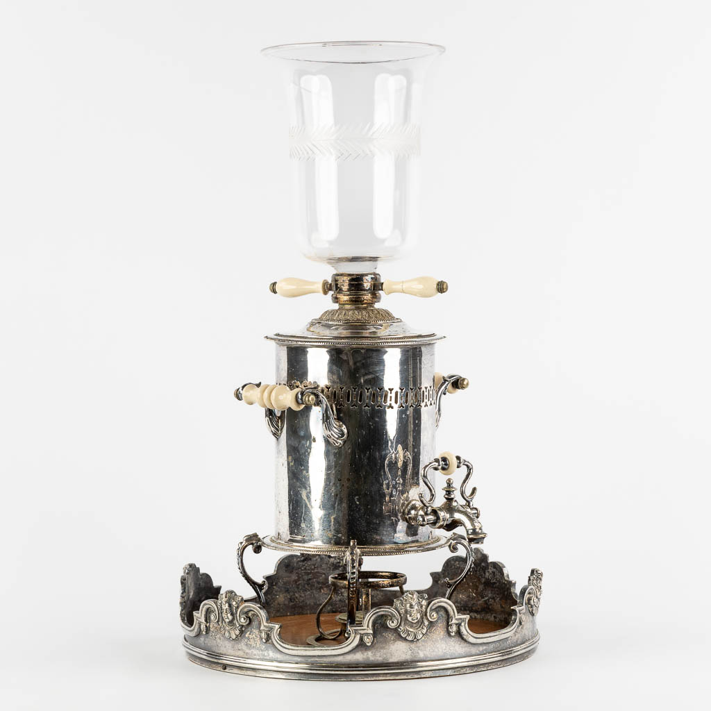 Een verzilverde samovar met glas, staande op leeuwen. (W:33 x H:51 cm)